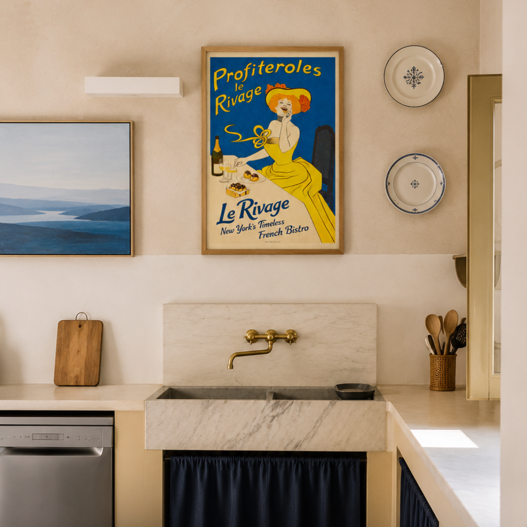 A styled kitchen interior featuring a vintage-style framed poster with the text 'Profiteroles Rivage' and 'Le Rouge' above a marble sink with a brass faucet. The scene includes a blue ocean painting, decorative plates, a wooden cutting board, and dark navy curtains under the sink.