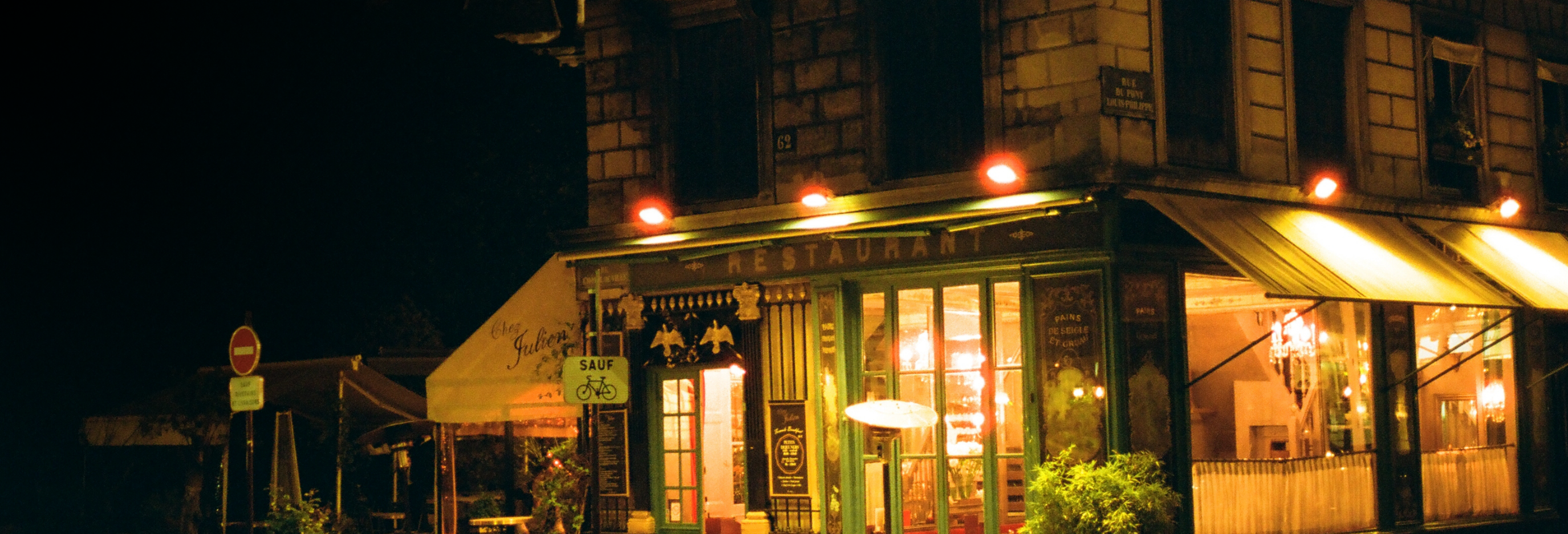 A nighttime street view of a Parisian-style cafe with outdoor seating, awnings, and warm yellow lighting glowing from the windows and above the entrance.