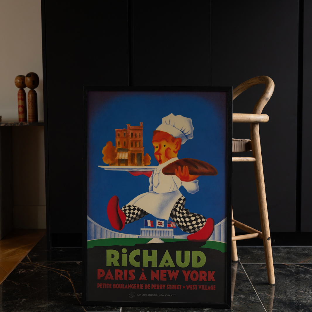 A framed vintage-style poster leaning against a dark cabinet on a marble floor, with a wooden chair beside it. The poster shows an illustrated chef carrying a tray with a pastry and the text 'RICHAUD PARIS A NEW YORK'.